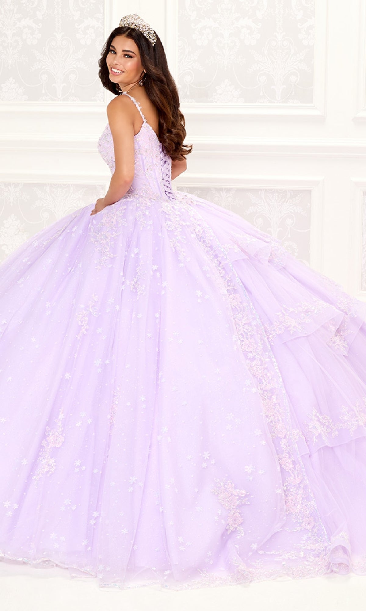 A woman wearing the Quinceanera Dress PR30087 By Princesa, featuring a lavender hue and corset bodice, stands in an ornate white room with a tiara, smiling over her shoulder.